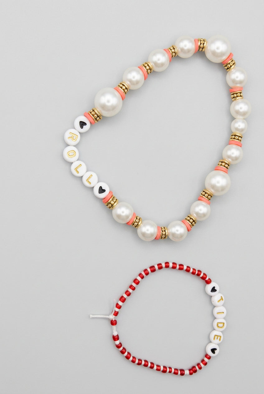 A beaded necklace and matching bracelet displayed on a textured beige fabric background. The necklace features white faux pearls, white letter beads spelling 'JUDY', and red and black spacer beads.