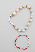 A beaded necklace and matching bracelet displayed on a textured beige fabric background. The necklace features white faux pearls, white letter beads spelling 'JUDY', and red and black spacer beads.