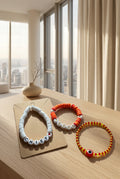 Three handmade beaded bracelets displayed on a kraft paper card against a textured beige fabric background. The bracelets include red and yellow evil eye beads, orange and white letter beads spelling 'LOVE', and white letter beads spelling 'CHIEF' with a small red heart.